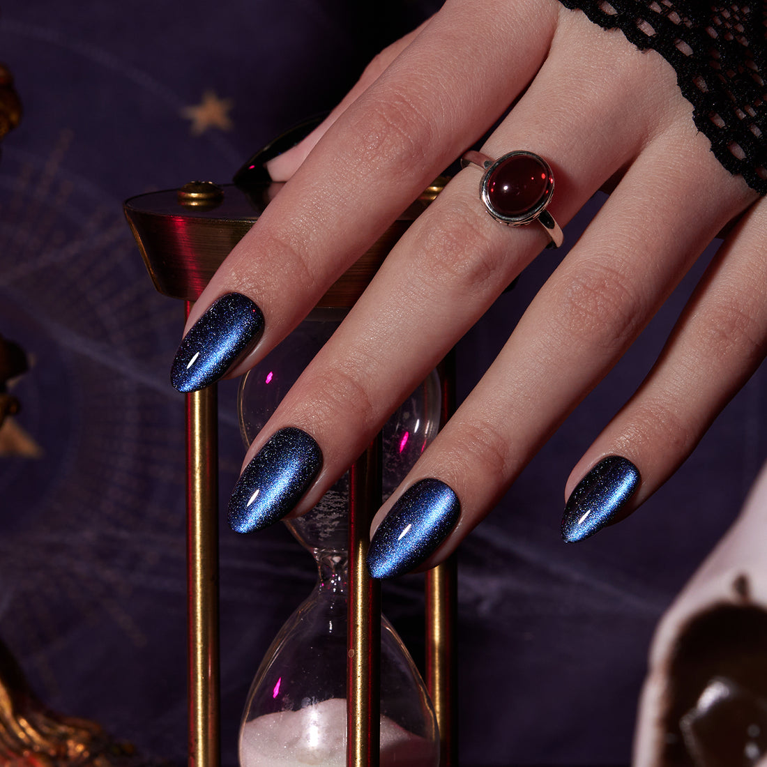 Model wearing deep blue magnetic cat-eye almond press-on nails beside an hourglass in a dark Halloween setting